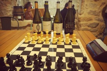 Chessboard ready for play on a wooden table with wine bottles and a rustic stone wall in the background.